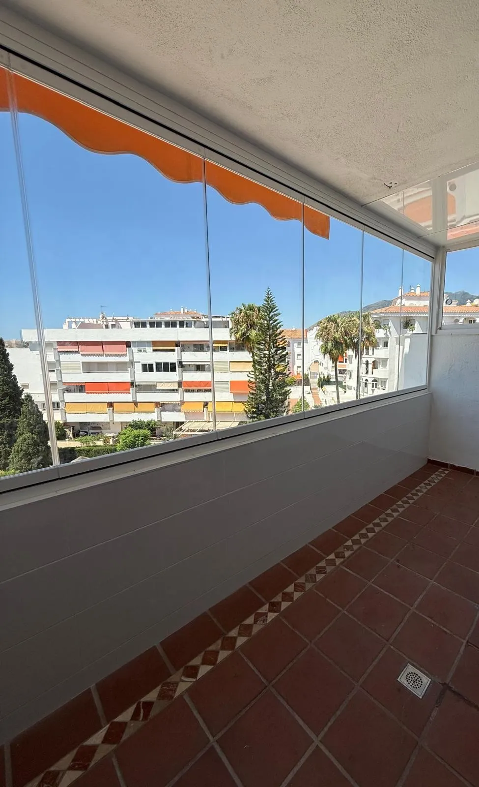 Terraza cerrada con cortina de vidrio sobre un murete blanco, bajo un toldo naranja