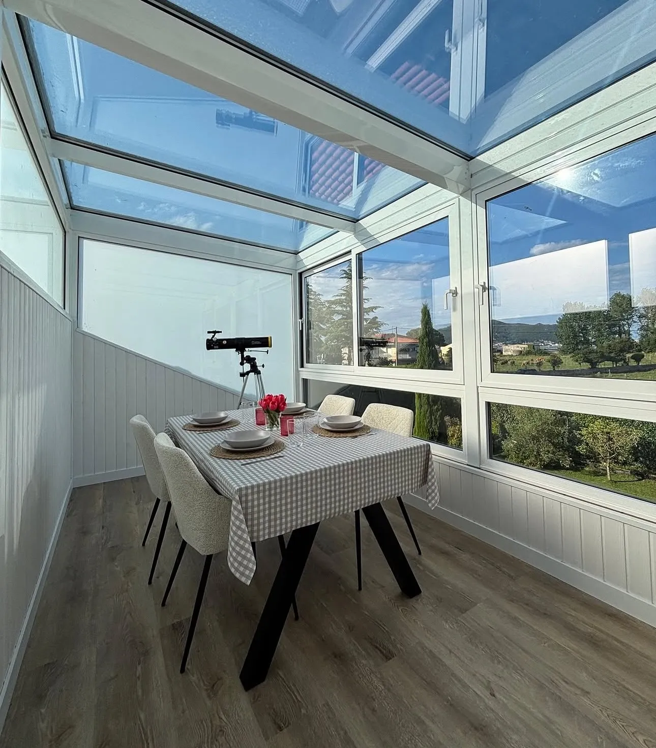 Interior de una galería acristalada de aluminio blanco con techo de cristal, mesa de comedor y vistas al campo