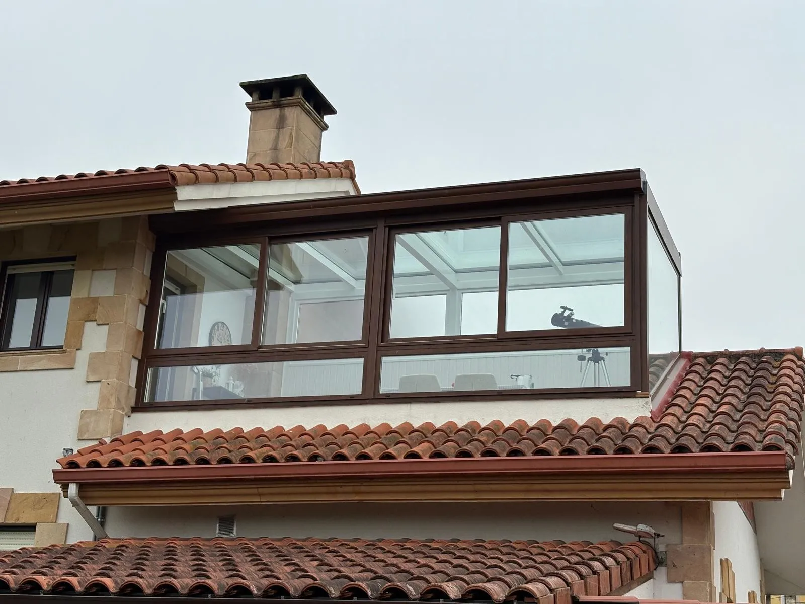 Galería acristalada con perfiles en tono madera levantada sobre el tejado de teja de una vivienda