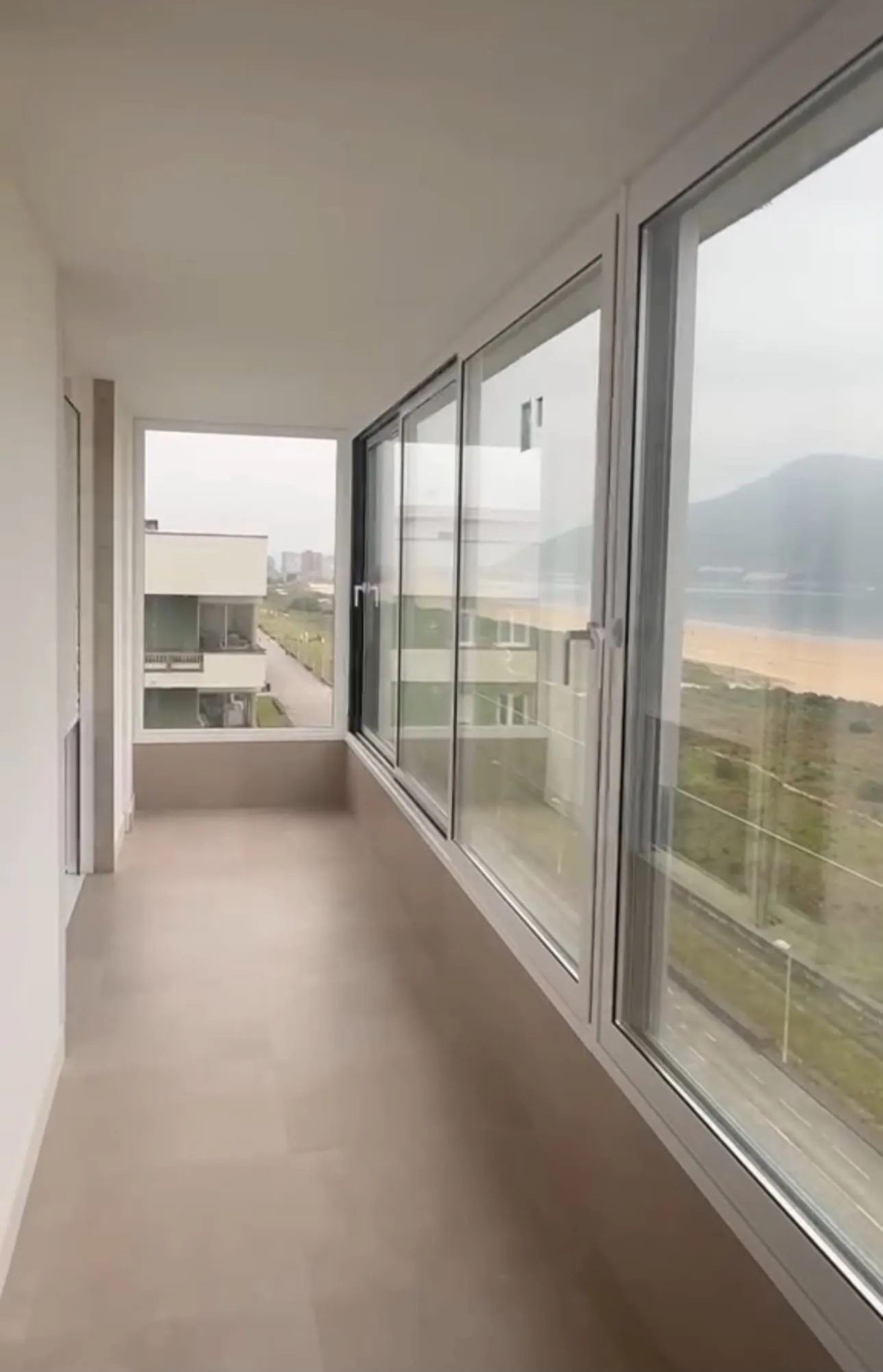 Terraza cerrada con ventanas correderas de aluminio blanco, con vistas a una playa y a las montañas