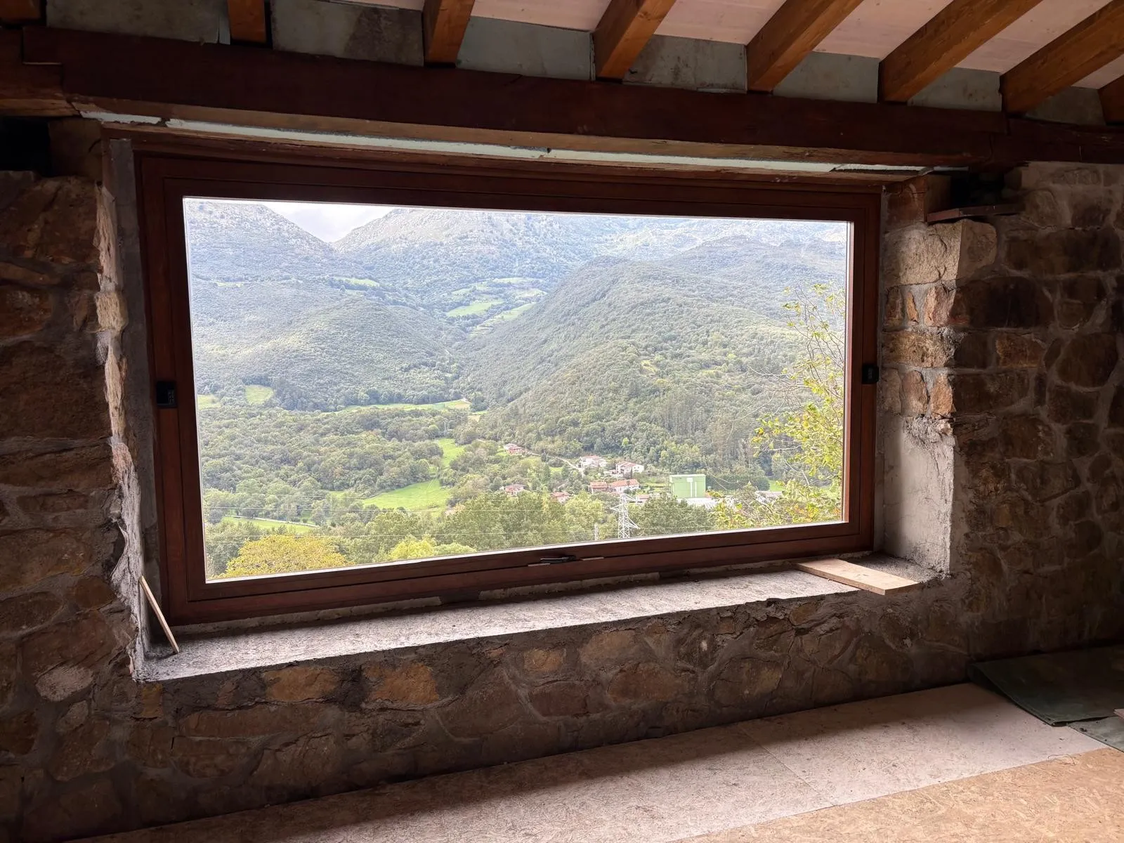Ventana panorámica con marco en tono madera en un muro de piedra bajo vigas de madera, abierta a un valle verde entre montañas