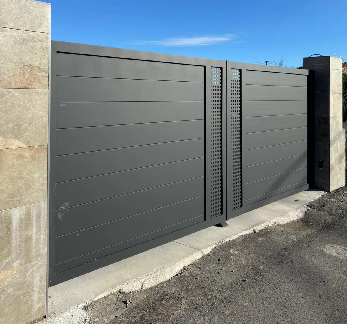 Puerta corredera de aluminio gris antracita con lamas horizontales y franjas perforadas, entre pilares de piedra