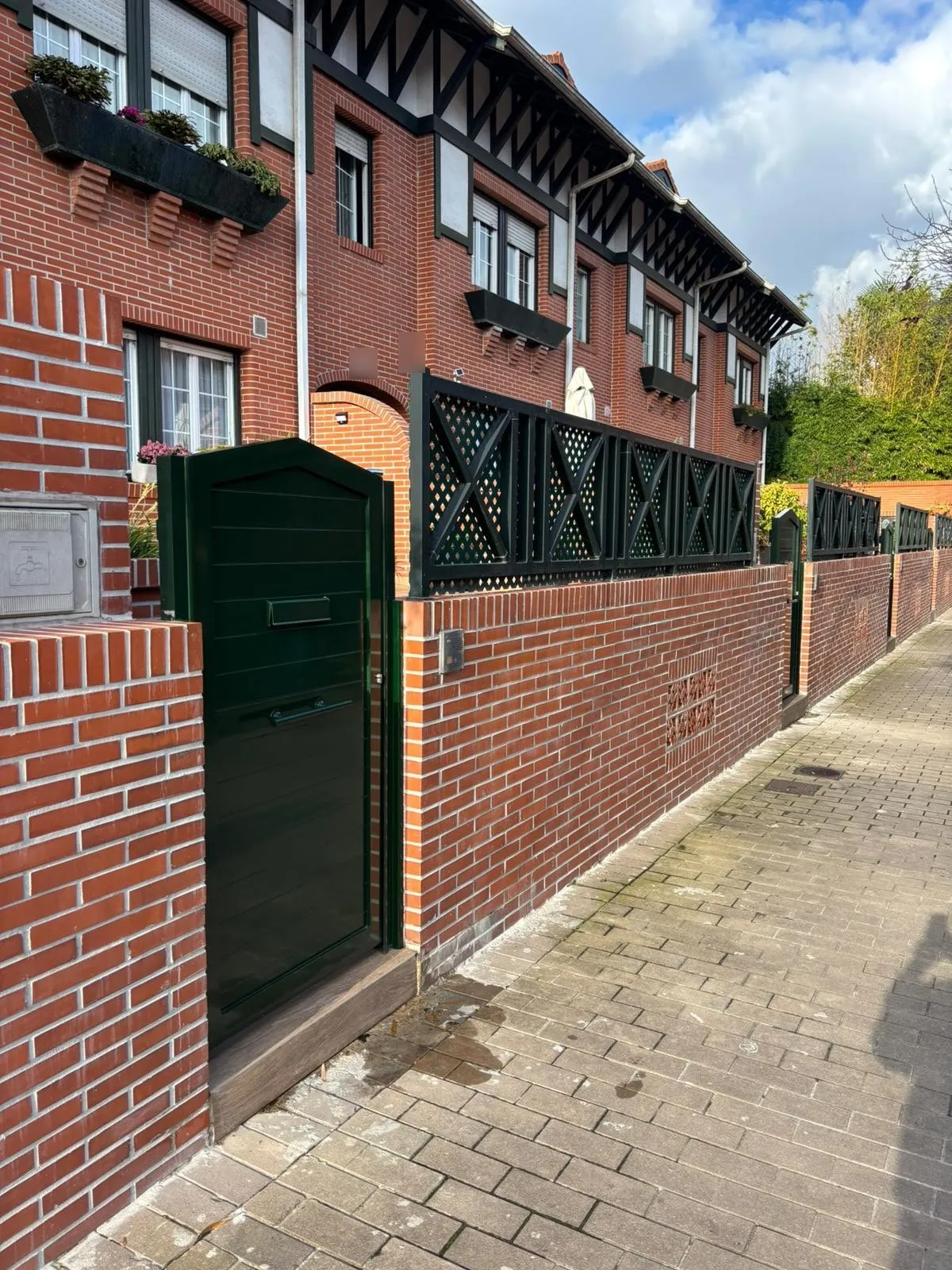 Puerta peatonal verde junto a un cerramiento de celosía verde sobre muro de ladrillo, a lo largo de la acera