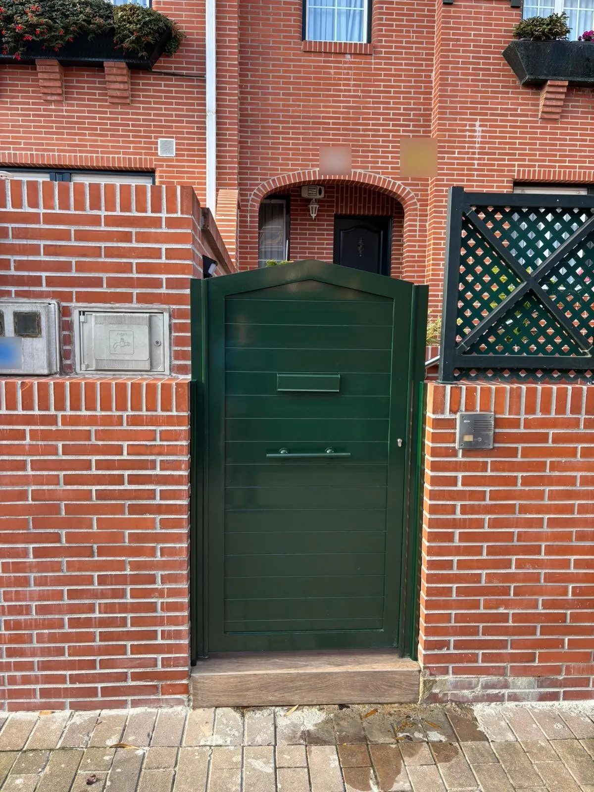Puerta peatonal de aluminio verde con lamas horizontales y buzón, en un muro de ladrillo caravista