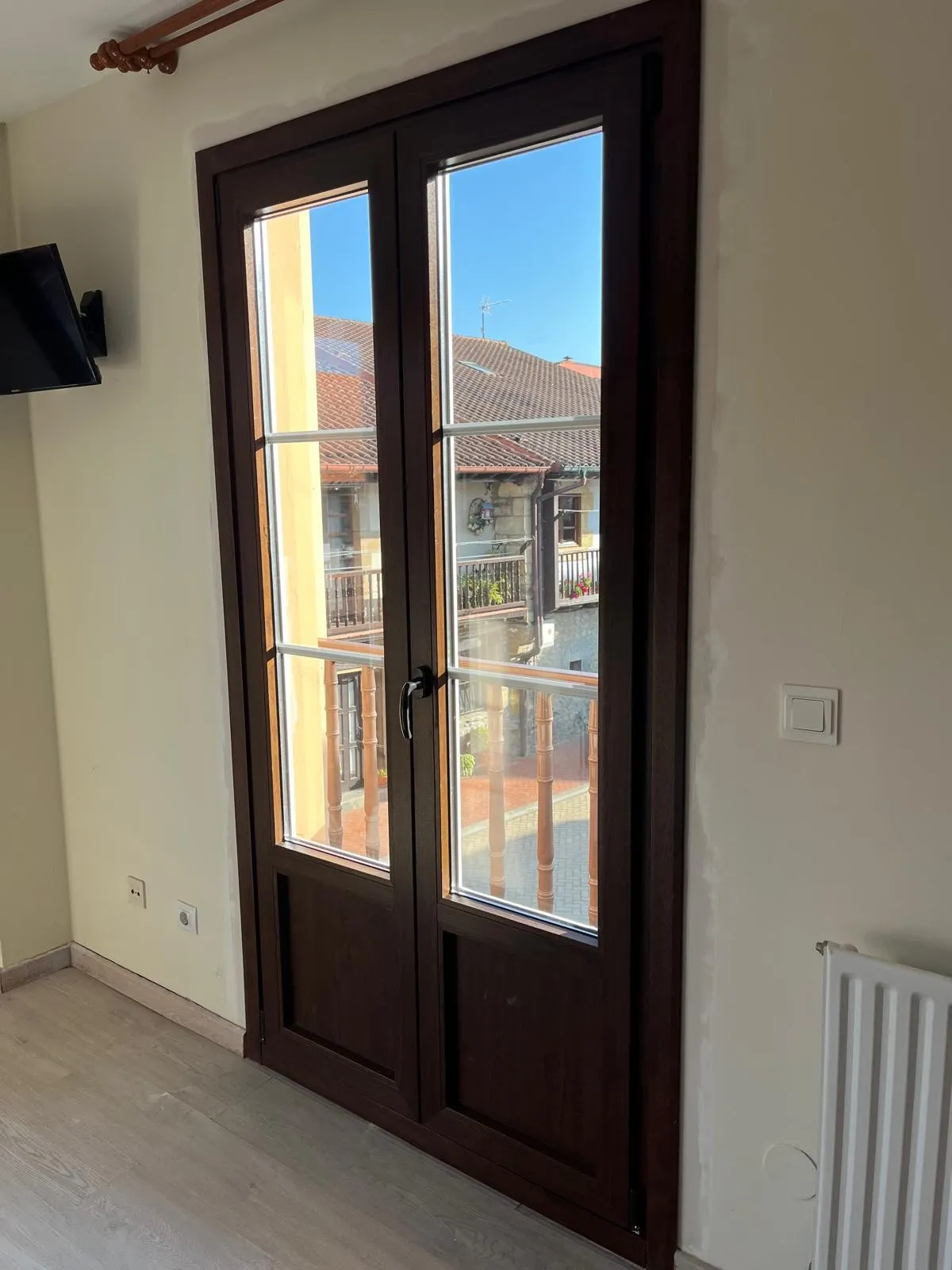 Puerta balconera de dos hojas en tono madera oscura, con cuarterones y zócalo, que da a un balcón de madera