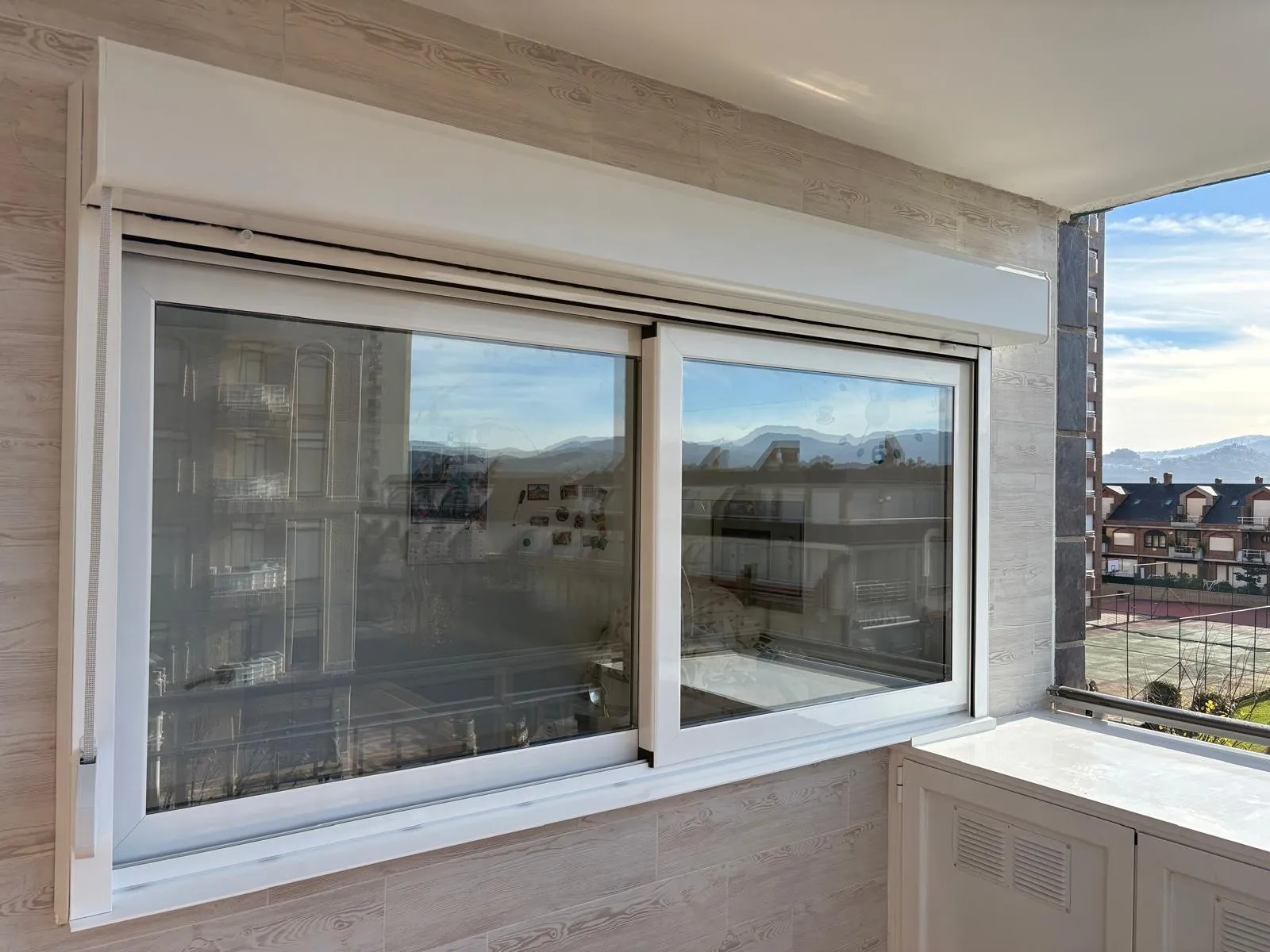 Ventana corredera de dos hojas en blanco con persiana enrollable, vista desde una terraza con montañas al fondo