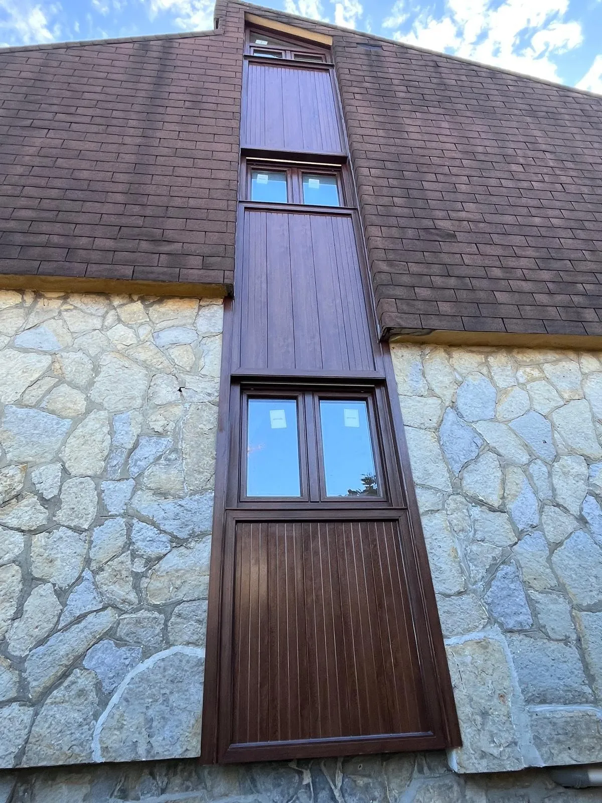 Conjunto vertical de ventanas y paneles en tono madera que recorre la fachada de piedra hasta el tejado