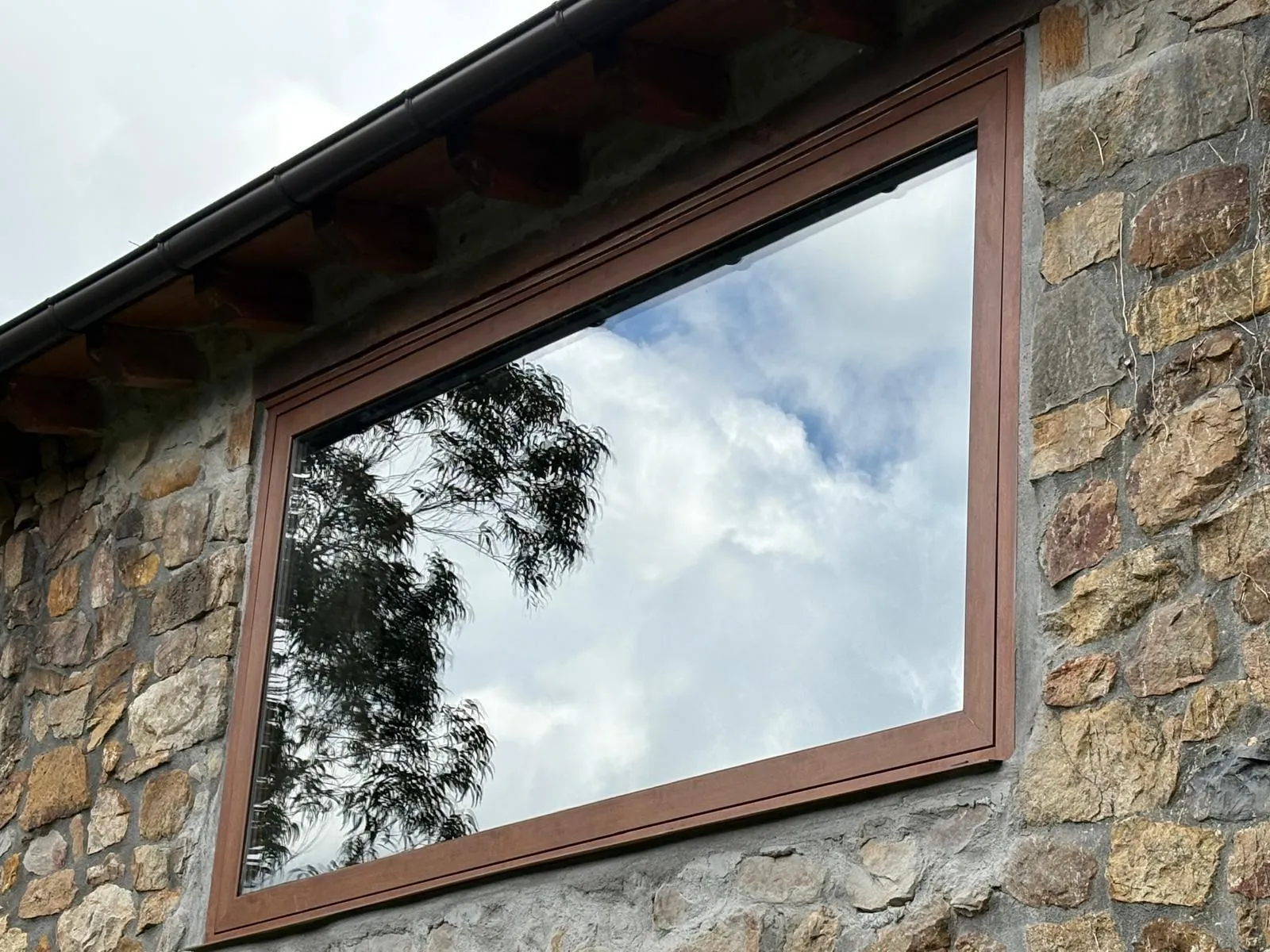 Ventana de gran tamaño con marco en tono madera en una fachada de piedra, con el cielo y un árbol reflejados en el vidrio